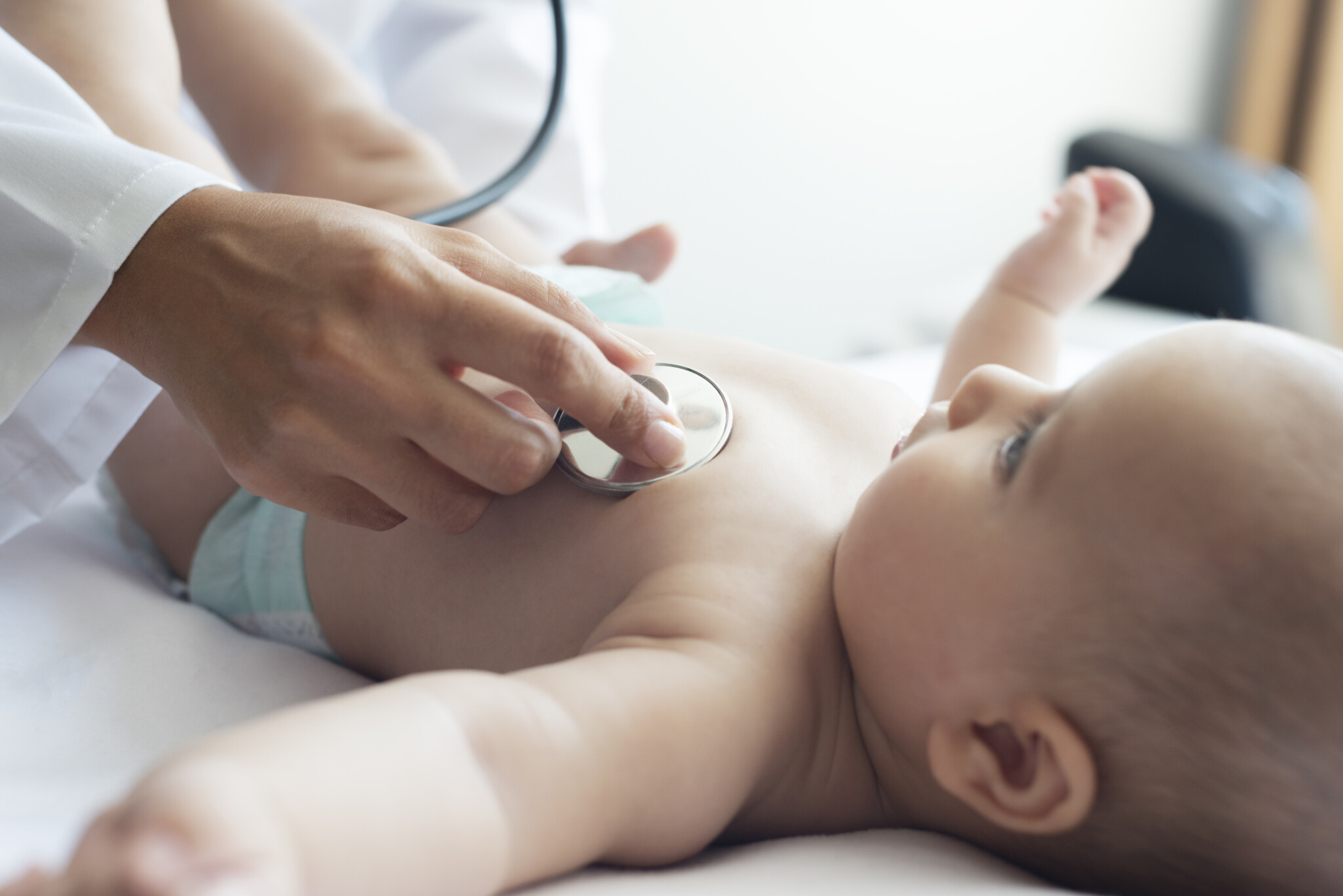 Baby lying on her back and is looking up to the pediatrician who is listening her heart.